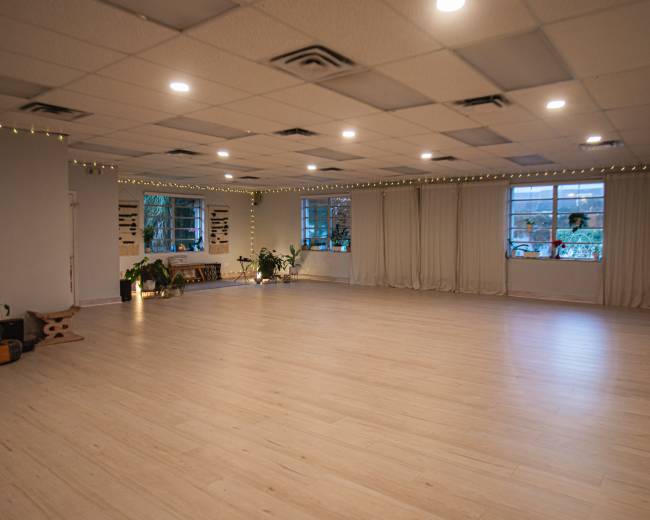 The image shows a spacious, well-lit room with wooden flooring, large windows, and plants along the walls.