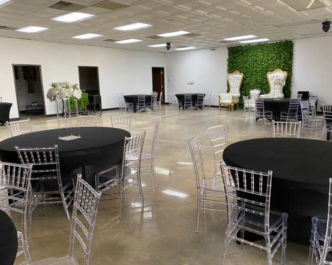 The image shows an indoor event space with black round tables and clear chairs, featuring a greenery wall and two ornate white chairs in the background.