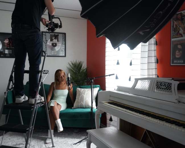 A camera operator on a ladder films a woman sitting on a green sofa in a room decorated with music memorabilia and a white piano.