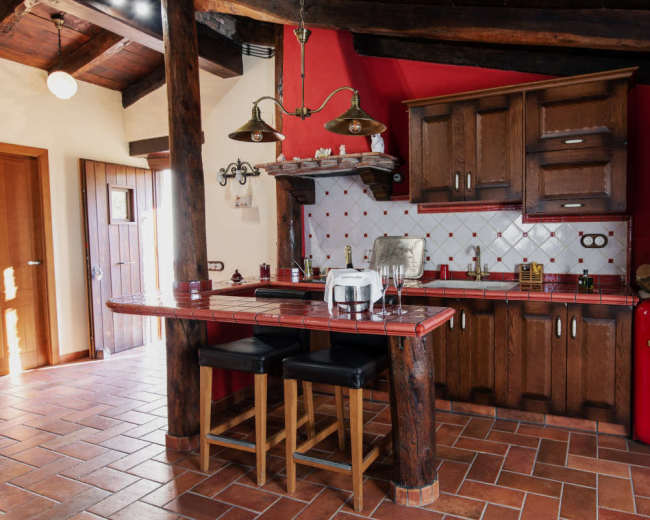 A rustic kitchen features wood cabinets, a red retro refrigerator, and a bar with two stools under hanging lights.