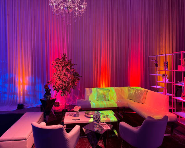 The scene features a modern lounge area with white furniture and colorful ambient lighting against a draped backdrop.