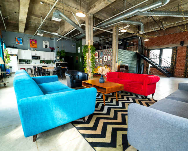 The image shows a spacious interior featuring a mix of colorful sofas, a wooden coffee table, and modern decor in an industrial-style loft.