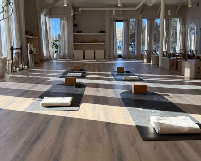 The image shows a spacious yoga studio with wooden flooring, multiple yoga mats arranged in rows, and large windows allowing natural light to illuminate the space.