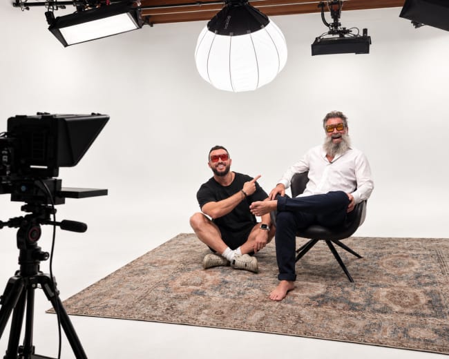 Two men sit in a studio setting on a patterned rug, with one man in a chair and the other seated on the floor, both wearing sunglasses and appearing relaxed.