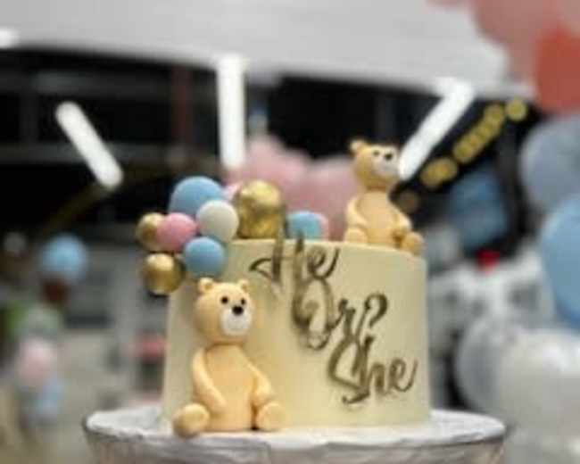 A two-tiered cake decorated with teddy bear figures and colorful balloon accents, featuring the text "He or She."