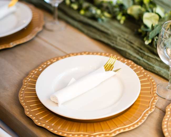 A dining table is elegantly set with white plates, golden chargers, wine glasses, and candles, accented by a green centerpiece.