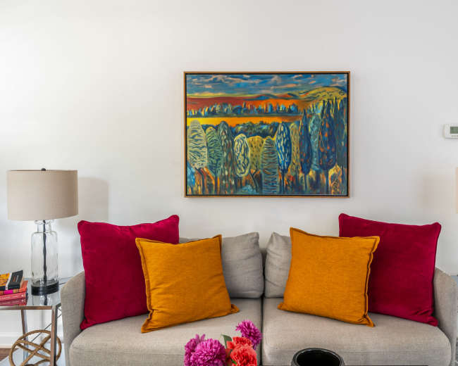 A gray sofa with orange and magenta pillows is positioned beneath a colorful landscape painting, flanked by clear table lamps on either side.