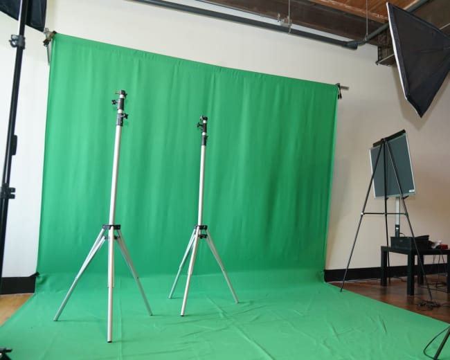 A photography studio setup featuring two light stands and a large green backdrop, with equipment and a stool visible in the background.