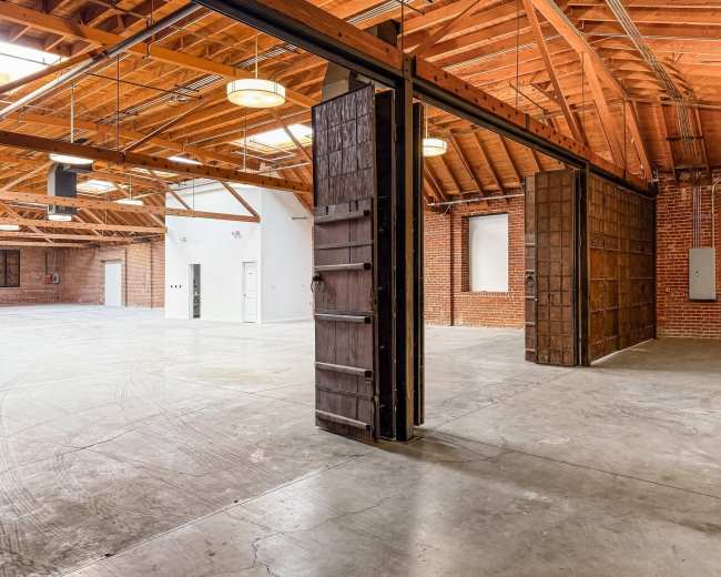 The image shows a spacious industrial interior with exposed wooden beams, brick walls, and large sliding doors.