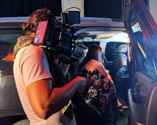 A camera operator films a woman sitting inside a vehicle, reviewing content on a device.