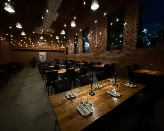 The image shows a spacious restaurant with wooden tables set for dining, illuminated by string lights and featuring exposed brick walls.