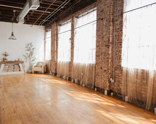 The image shows a spacious, well-lit interior with wooden floors, large windows adorned with sheer curtains, and minimal furniture, including a sofa and a decorative table.