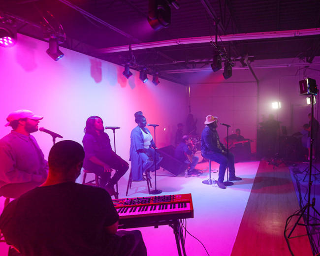 A group of musicians performs on stage under colorful lighting while a technician operates equipment nearby.