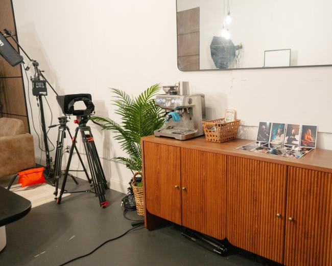 The image shows a wooden sideboard with a coffee machine and magazines, set against a plain wall, alongside a camera equipment tripod and a potted plant.
