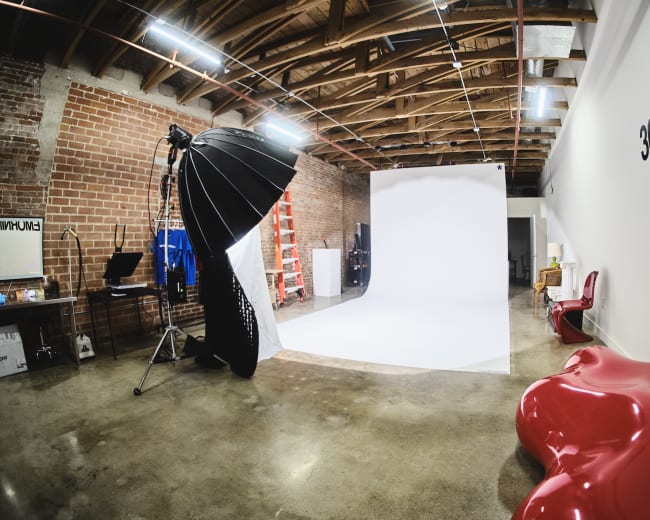 The image shows a photography studio with a white backdrop, lighting equipment, a shiny red sculptural chair, and exposed brick walls.