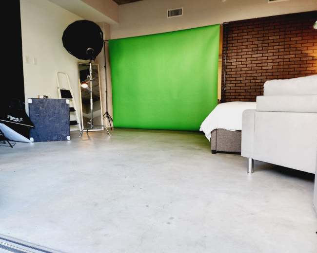 The image shows a studio space featuring a green backdrop, a brown brick wall, a white sofa, and a few lighting equipment pieces.