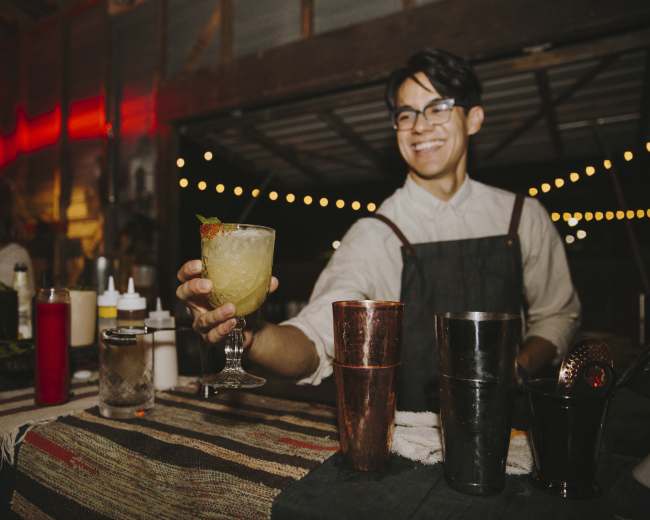 A bartender in an apron smiles while holding a cocktail at a well-lit bar in an outdoor setting.