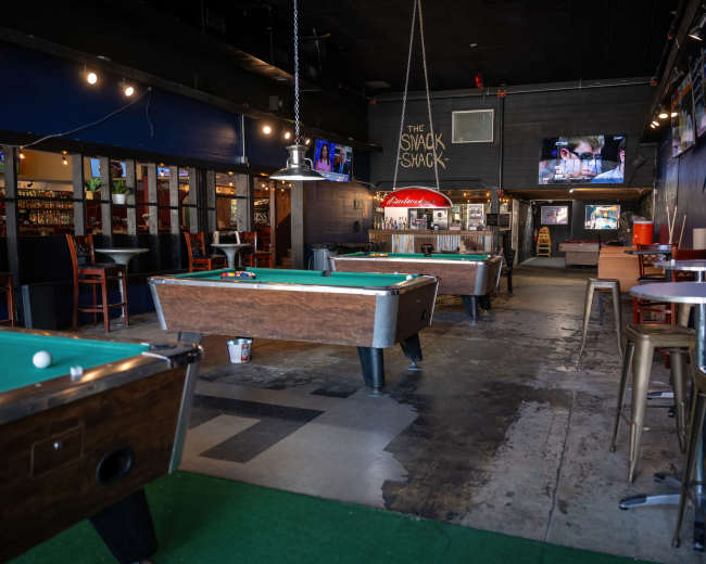 The interior of a casual bar features several pool tables, a bar area in the background, and a mix of seating arrangements.