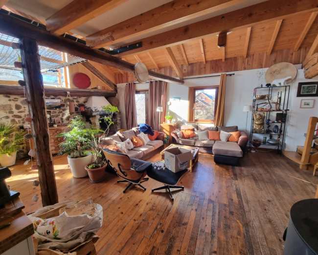 A spacious living room features a large sectional sofa with colorful cushions, wooden beams, and plants, along with a mix of wooden and modern furniture.