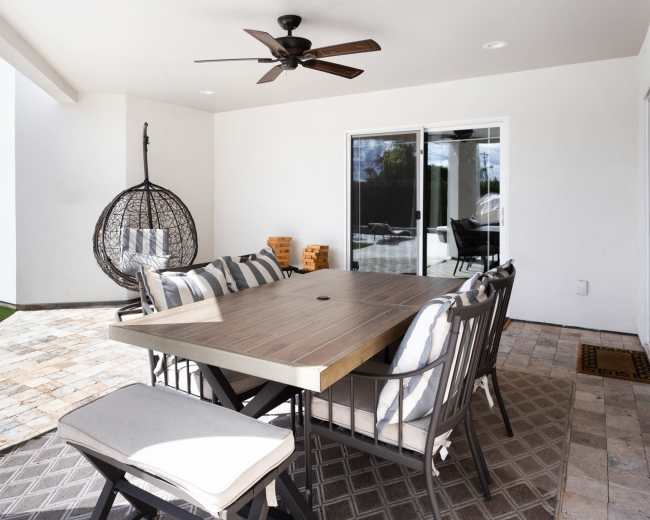 The image shows a patio area with a large wooden dining table, several chairs, a hanging chair, and a ceiling fan, all set against a backdrop of white walls.
