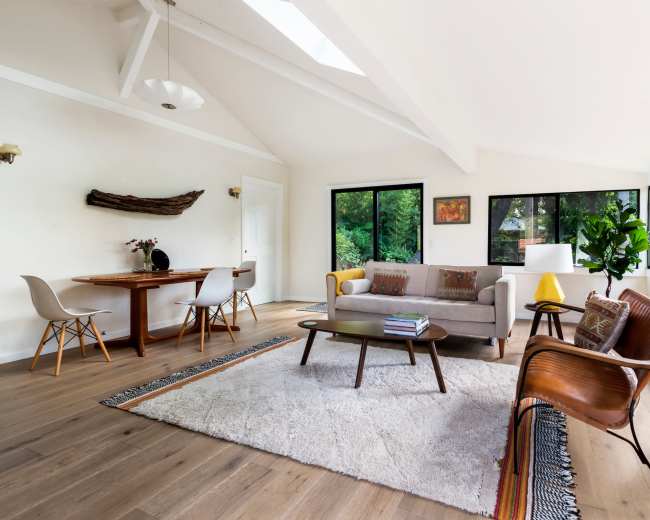 A bright living room with a beige sofa, wooden furniture, and large windows overlooking greenery.