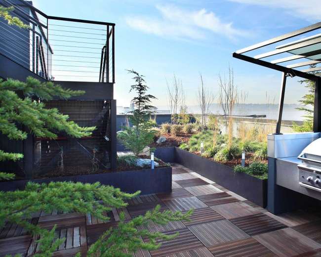 A rooftop terrace featuring a stainless steel barbecue grill, landscaped planters, and a view of the sky and distant landscape.