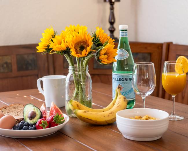 A breakfast table is set with two eggs, a bowl of cereal, assorted fruits, a glass of orange juice, sparkling water, and a vase of sunflowers.