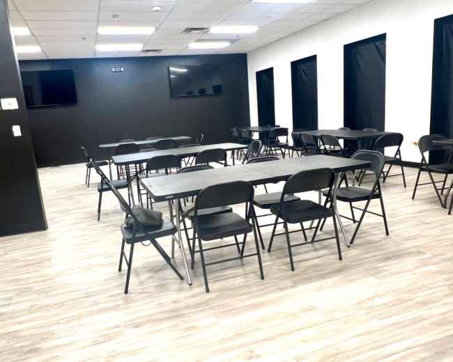 The image shows a minimalist meeting room with several folding tables and chairs arranged in a grid pattern, along with two wall-mounted screens.