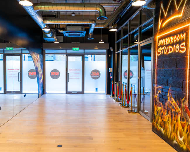The image shows a spacious studio entrance with glass doors, wooden flooring, and a wall featuring neon text.