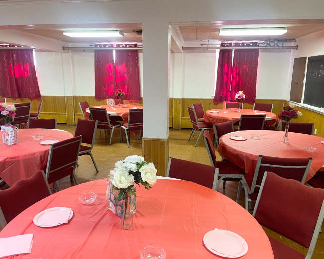 The image shows a banquet hall with round tables covered in orange tablecloths, each set with plates and flower centerpieces, and red curtains framing the windows.