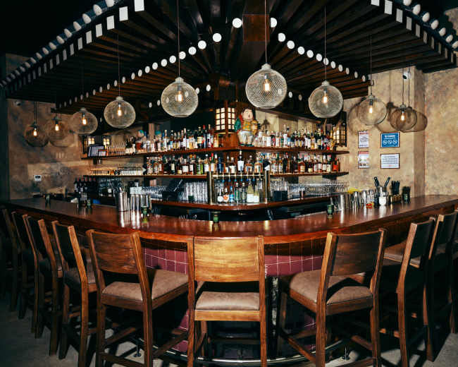 The image shows a curved wooden bar with high stools, surrounded by shelves filled with various bottles and decorative light fixtures overhead.