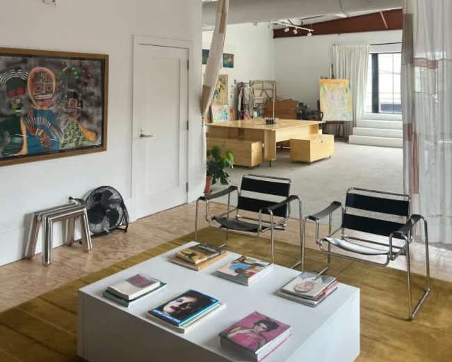 The image shows a spacious, modern room with a seating area featuring two striped chairs and a coffee table displaying books, adjacent to an open area with art supplies and artworks.