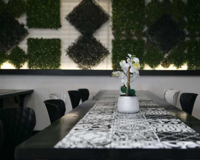 A long dining table with a patterned table runner is set in a restaurant, surrounded by black and white chairs, with decorative plants on the wall behind.
