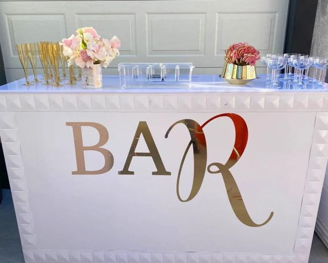 A bar setup featuring a white counter with a gold "BAR" sign, adorned with floral arrangements, glassware, and cocktail supplies.