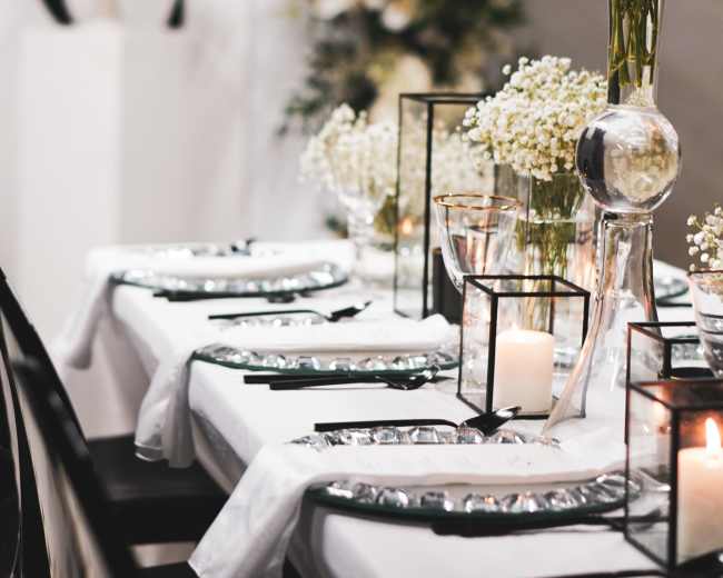 A elegantly set dining table features white tablecloths, glass vases with flowers, candles, and decorative accents in a styled venue.