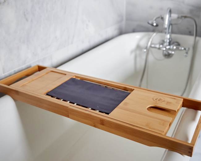 A wooden bath tray rests across the edge of a white tub in a marble-tiled bathroom.