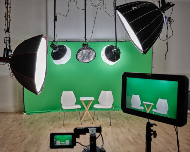 A studio setup features two white chairs facing a small table in front of a green backdrop, illuminated by five overhead lights and monitored by a camera.