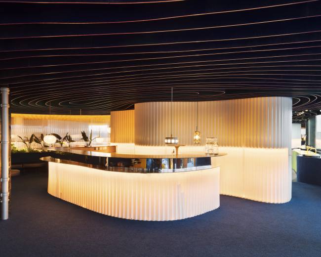 A modern bar area with a curved light-colored counter, featuring a black bar top and illuminated by warm lighting against a backdrop of dark, wavy ceiling details.