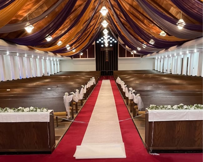 The image shows a decorated interior of a church with wooden pews along the sides, a red carpet down the aisle, and a floral arrangement at the front.