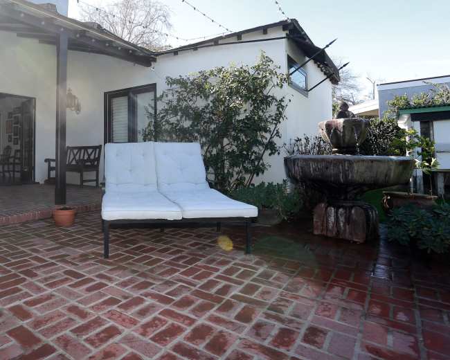The image shows a patio area featuring a white, cushioned lounge chair beside a stone fountain and surrounded by greenery.