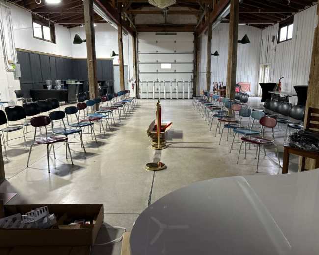 A spacious room with a high ceiling is set up with rows of chairs facing a central podium, flanked by tables and large garage doors at one end.