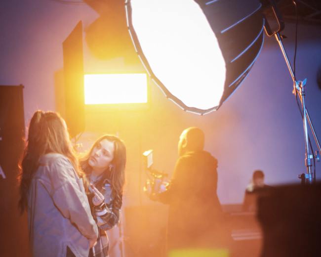The image shows two women in a studio setting illuminated by a large light source, with soft fog in the air and candles arranged on the floor.