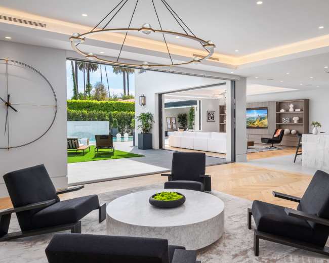 The image shows a modern living space featuring a circular coffee table surrounded by black lounge chairs, with an open view to an outdoor area and a kitchen in the background.