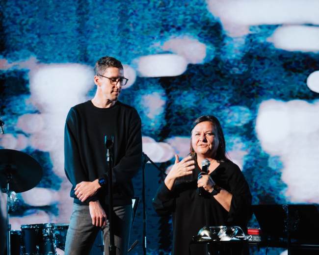 A man and a woman are speaking on stage in front of a large screen, with musical instruments visible in the background.