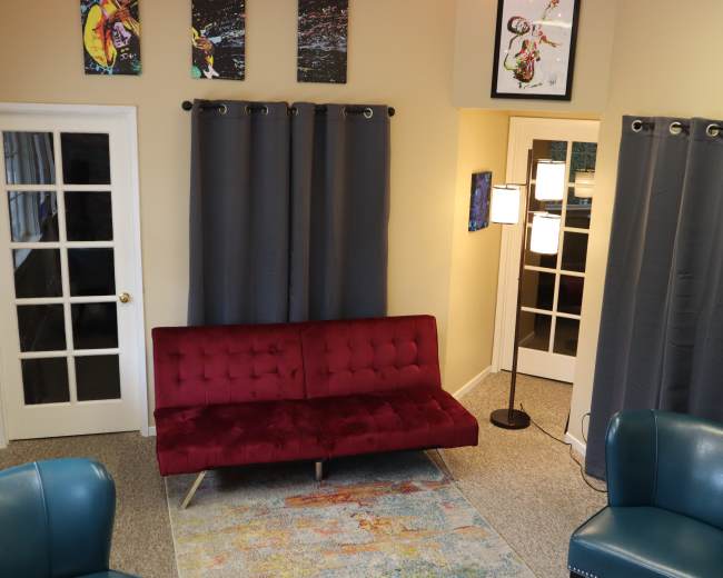 The room features a red velvet couch, two teal chairs, gray curtains, and artwork on the walls, with a floor lamp in one corner.