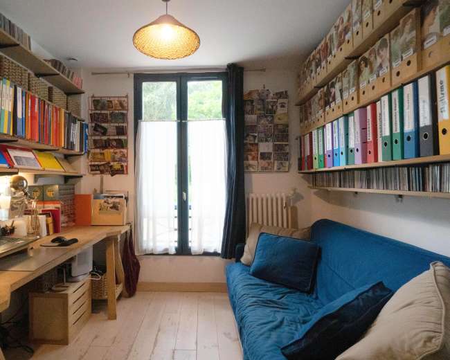 A home office features a blue sofa, a desk with a computer, and shelves filled with colorful binders and photo frames.