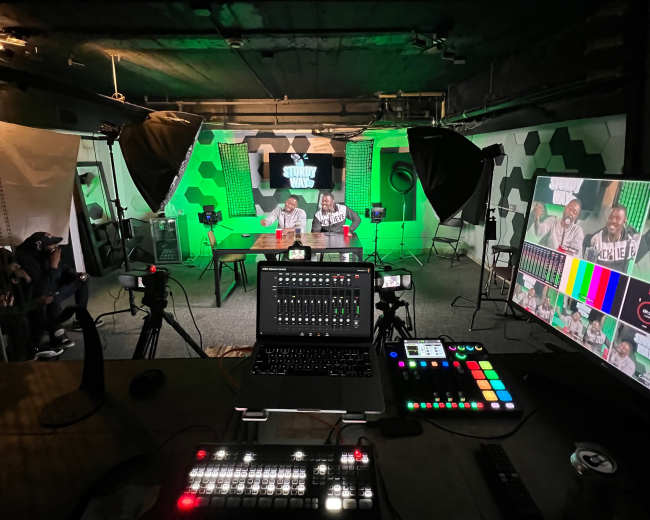 A recording studio setup with two people seated at a table under green lighting, surrounded by cameras, monitors, and production equipment.