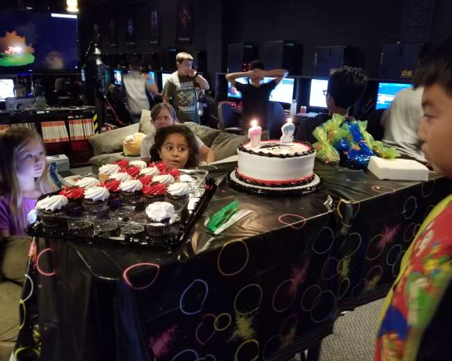 A birthday celebration takes place in a gaming lounge with a table set up featuring a decorated cake and cupcakes, surrounded by children and gaming monitors in the background.
