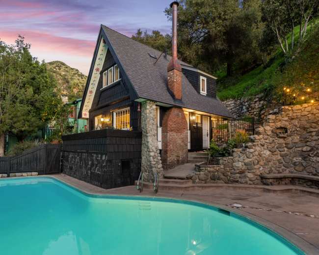 The image shows a charming house near a swimming pool, surrounded by trees and rocky terrain, with a scenic mountainous backdrop at dusk.
