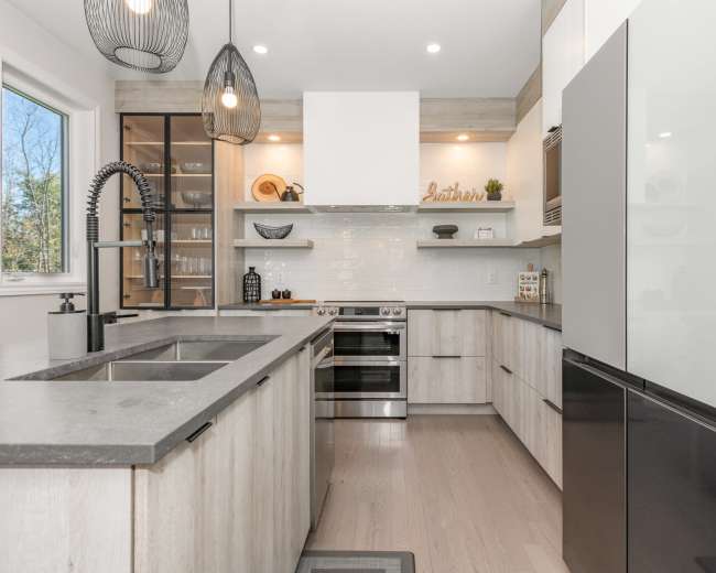 A modern kitchen features light wood cabinetry, a gray countertop with a double sink, and stainless steel appliances.